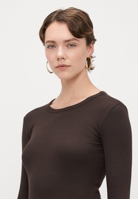 Camisola de manga longa castanha com decote redondo, apresentando um corte justo que acentua a silhueta do corpo. Sem padrões ou texturas visíveis.