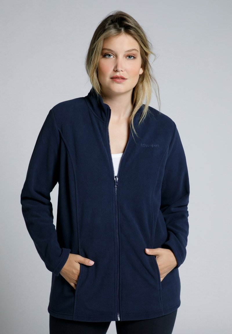 Tummansininen fleece-takki, jossa on vetoketjusuljenta, pystykotelo ja sivutaskut. Pehmeä rakenne ja muotoinen malli, joka sopii arkivaatteeksi.