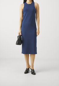 Kobieta w granatowej, bezrękawnikowej sukience midi, trzymająca małą czarną torebkę i nosząca czarne, na obcasie klapki na jednolitym tle.