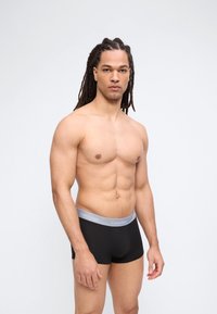 Μυώδης άνδρας με μακριά dreadlocks που φοράει μαύρα εσώρουχα Calvin Klein boxer briefs σε ουδέτερο ανοιχτό φόντο.