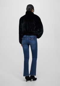 Kvinde iført en sort faux fur jakke, blå flagrende jeans med baglommer og sorte højhælede ankelstøvler, stående med ryggen til.