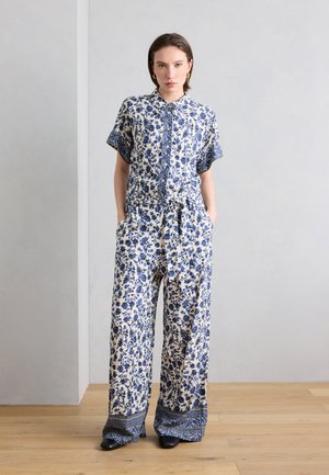 Naine seisab siseruumis, kandes sinise ja valge lillemustriga lühikeste varrukatega nööpidega särki ning sobivaid laiade säärtega pükse mustade kingadega.