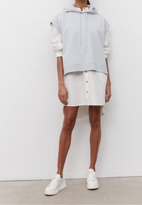 Světle modrá zkrácená mikina s klokaní kapsou a stahovací šňůrkou, vrstvená přes bílou košilovou šaty s vysokým a nízkým lemem, doplněná bílými teniskami.