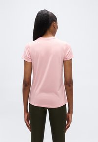 Γυναίκα με σκουρόχρω μη αλογοουρά που φοράει ανοιχτό ροζ μπλουζάκι με κοντά μανίκια και σκούρο πράσινο παντελόνι, στέκεται γυρισμένη με την πλάτη προς το γυαλιστερό φόντο.