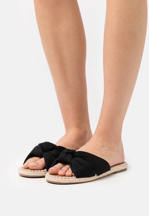 Piedi che indossano sandali slip-on con nodo in tessuto nero e suole intrecciate beige su uno sfondo bianco.