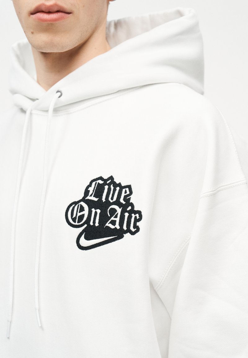 Teilansicht einer Person, die einen weißen Kapuzenpullover mit schwarzem "Live On Air"-Text und Nike-Swoosh auf der Brust trägt.
