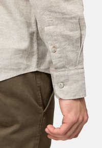 Stribet beige skjorte med en knapmanchet med kontrastfarvet orange tråd, parret med olivengrønne bukser. Tekstiler.