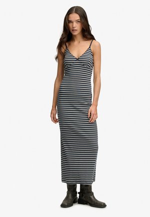 Kobieta ubrana w czarno-białą, poziomo paskowaną, bezrękawną sukienkę maxi z cienkimi ramiączkami i czarne botki za kostkę, stojąca na białym tle.