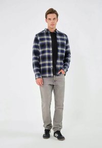 Kariertes Hemd in Marineblau und Beige, mit Knopfleiste, langen Ärmeln, über einem schwarzen Shirt getragen, kombiniert mit hellgrauen Jeans und dunklen Sneakers.