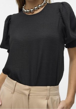 Femme portant un haut noir à manches bouffantes, pantalon beige taille haute, et un grand collier de perles argentées, la main reposant près de la poche.