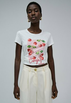 Camiseta de corte cropped de algodón blanco con un gráfico de cereza en rojo y verde, con el texto "Cerejas" debajo en rojo, acompañado de unos pantalones de talle alto beige.