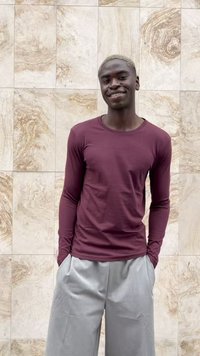 Smiling young man with short blond hair wearing a maroon long-sleeve shirt and light gray pants standing against a stone tile wall.