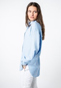 Lichtblauw, gestructureerd overhemd met lange mouwen, relaxed fit en afgeronde zoom, gecombineerd met witte broek. Model poseerde zijwaarts.