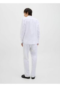 Camisa blanca de manga larga con cuello abotonado, de tejido ligeramente texturizado y con un plisado en la parte posterior, combinada con pantalones blancos a juego y sandalias negras.