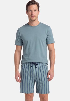 Man draagt een lichtblauw shirt met korte mouwen en blauwe gestreepte short met trekkoord, staand tegen een eenvoudige lichte achtergrond.