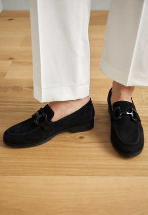 Persona con pantalones blancos recortados y mocasines de ante negro con una hebilla decorativa negra, de pie sobre un suelo de madera clara.