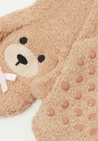 Pantofole marroni pelose con faccia d'orso, con caratteristiche ricamate e un fiocco rosa. La suola ha punti di gomma rotondi e texturizzati per una presa migliore.
