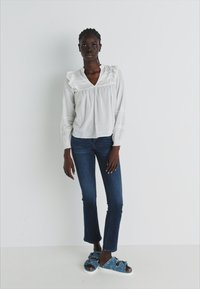 Vit blus med volangkrage och långa ärmar, i kombination med mörkblå skinny jeans och blå slitna slidesandaler.