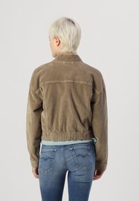 Brun kordfløyelsjakke med krage, elastisk midje og lange ermer. Brukt over en lyseblå topp, kombinert med blå jeans som har baklommer.