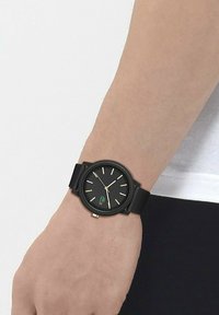 Montre noire avec un cadran noir texturé, des index des heures dorés et un logo vert. Elle dispose d'un bracelet en maille noire et a une forme ronde.