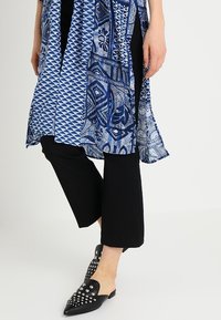 Kobieta w czarnych, krótkich spodniach, czarnych butach slip-on z ćwiekami oraz w zwiewnej, niebieskiej tunice w geometryczne i kwiatowe wzory.