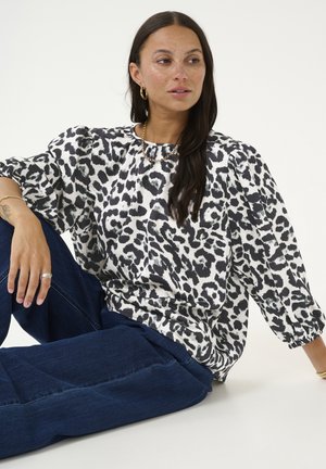 Femme aux longs cheveux foncés portant un chemisier imprimé léopard noir et blanc et un jean bleu, assise par terre avec une expression neutre.