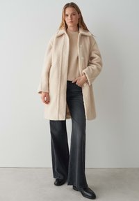 Cappotto in pelliccia ecologica beige con colletto, tre bottoni e tasche anteriori, abbinato a un maglione chiaro e jeans svasati scuri. Scarpe nere.