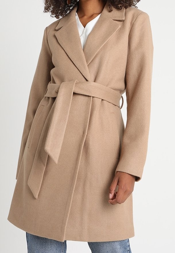 Femme portant un manteau en laine beige mi-genoux avec une ceinture nouée, une chemise blanche en dessous et un jean bleu sur un fond uni.
