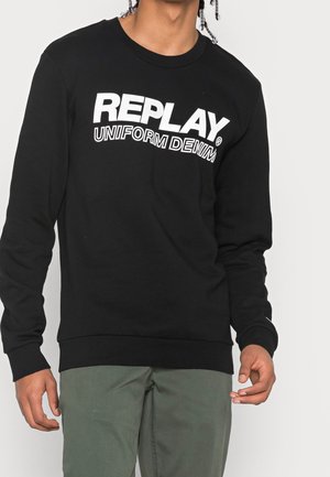 Mężczyzna w czarnej, długorękawowej bluzie z białym napisem "REPLAY UNIFORM DENIM", w połączeniu z oliwkowozielonymi spodniami, na jednolitym tle.