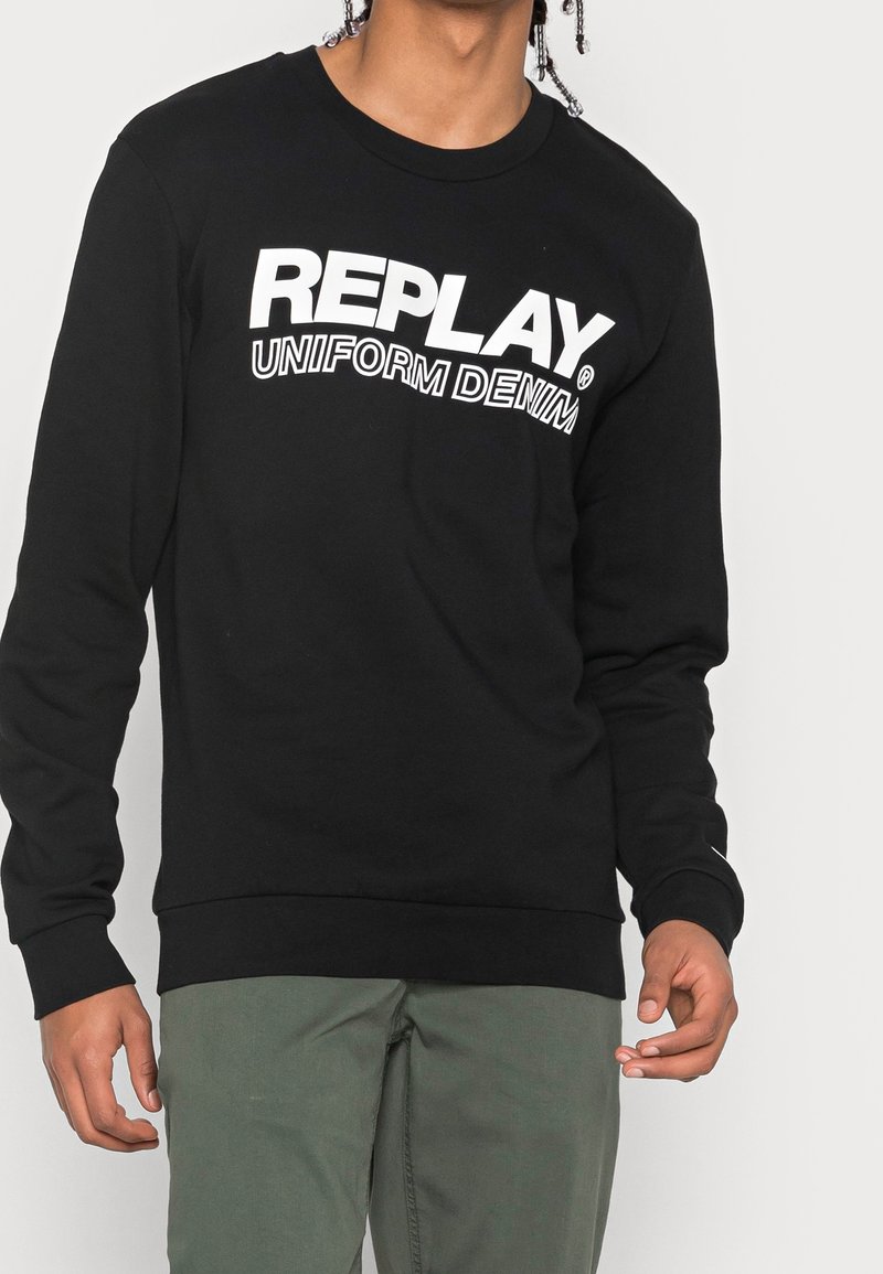 Homme portant un sweat-shirt à manches longues noir avec le texte blanc « REPLAY UNIFORM DENIM », associé à un pantalon vert olive, sur un fond uni.