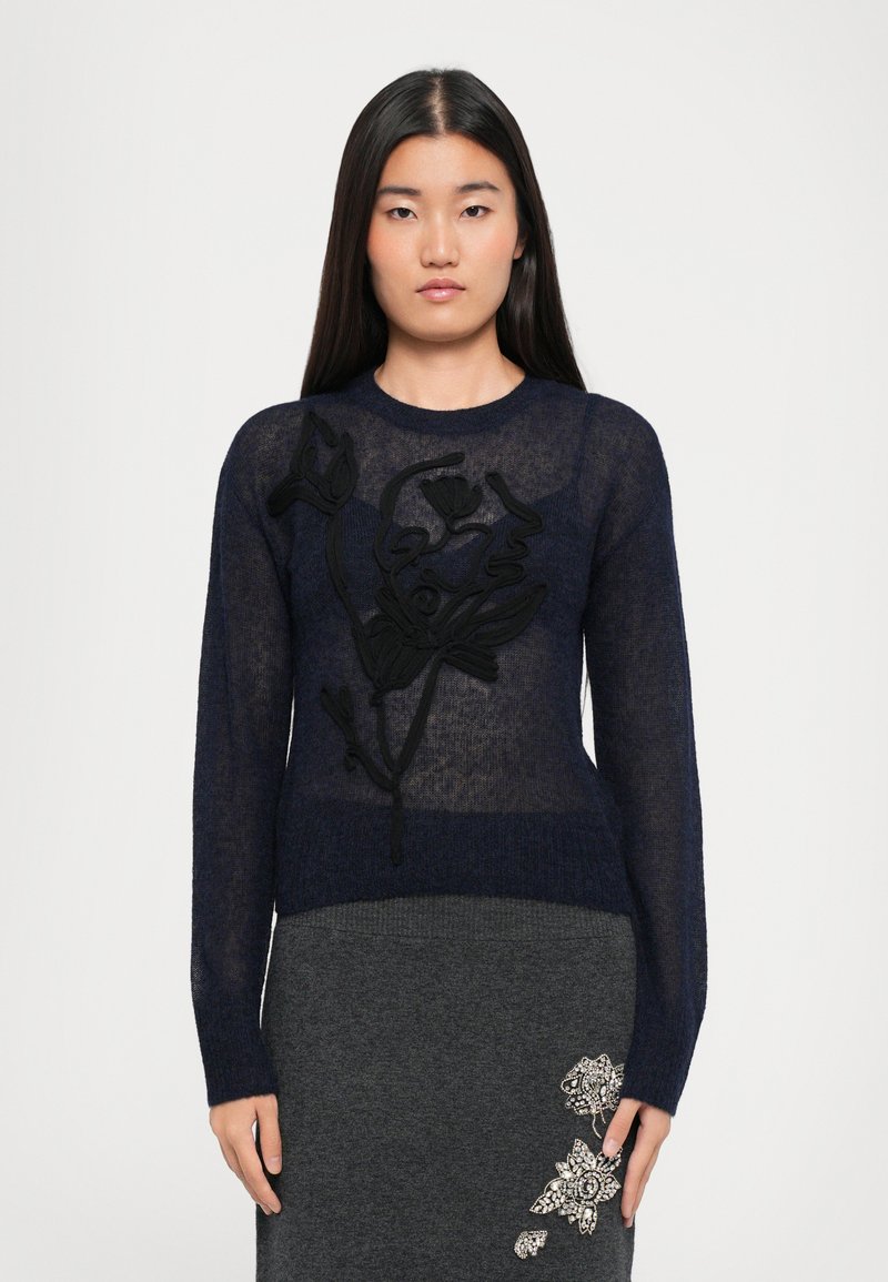Maglione trasparente blu navy con maniche lunghe, caratterizzato da ricami floreali neri sul davanti, abbinato a una gonna grigia decorata con dettagli.
