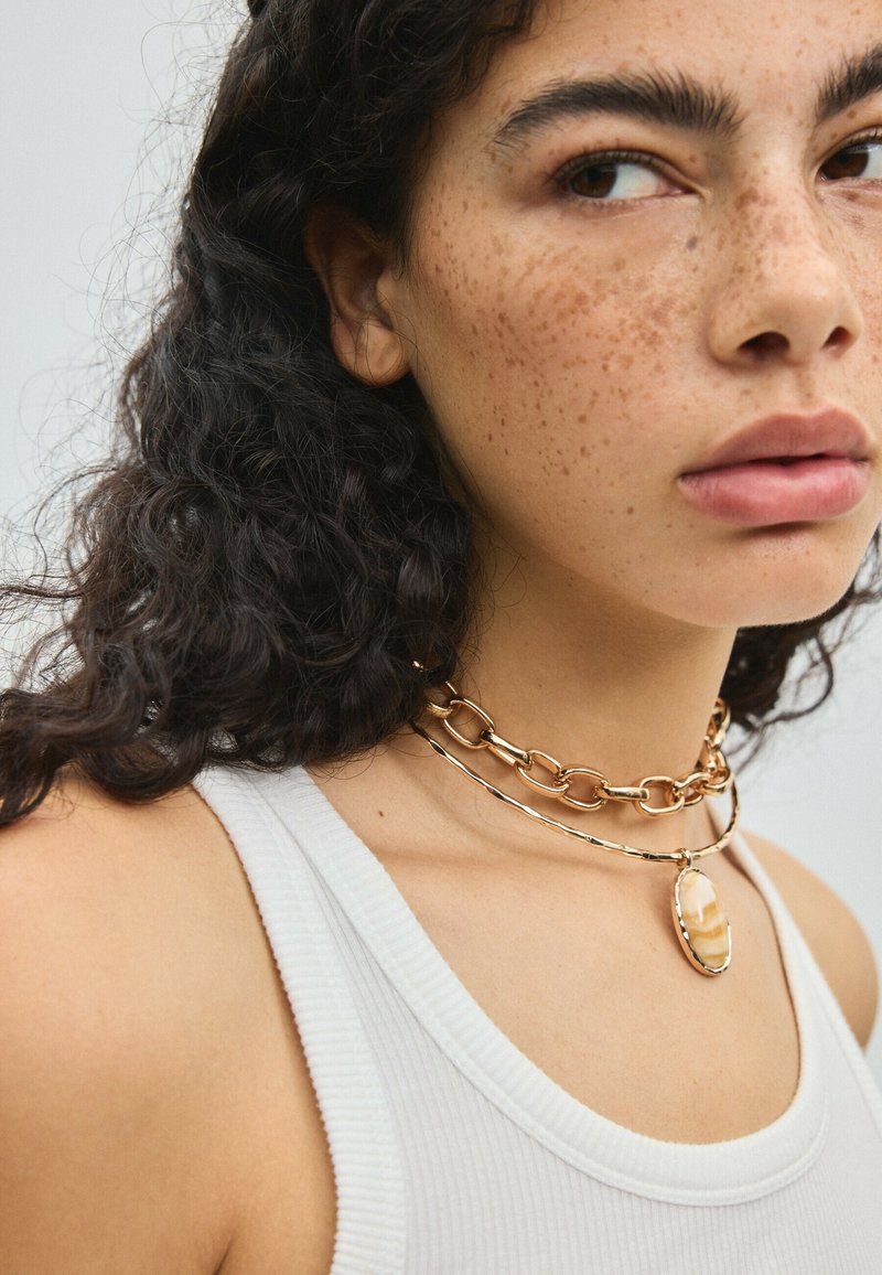 Femme aux cheveux bouclés foncés et taches de rousseur portant un débardeur blanc côtelé et deux colliers en or, dont un avec un grand pendentif ovale.