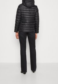 Veste doudoune noire isolée avec capuche, présentant un design matelassé et un jean noir évasé. Le tissu semble lisse et brillant.