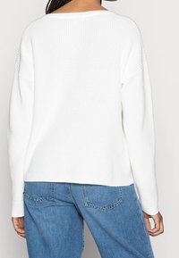 Witte gebreide trui met een wijde halslijn, verlaagde schouders en geribde textuur. Gepaard met lichtblauwe jeans die de naden van de achterzakken laten zien.