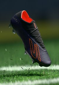 Scarpino da calcio nero con superficie texturizzata, accenti arancioni vivaci e dettagli di allacciatura, sospeso sopra erba verde e terra sparsa.