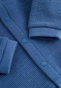 Close-up of blue waffle-knit fabric with ribbed cuffs and matching blue snap buttons on a garment.
