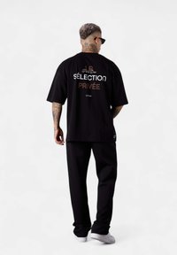 Schwarzes, oversized T-Shirt mit weißem und metallischem Schriftzug auf dem Rücken, kombiniert mit lockeren schwarzen Hosen und weißen Sneakers. Flaches, glattes Material.