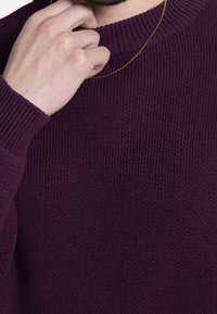 Mørkelilla strikket sweater med ribbet struktur, med rund halsudskæring og synlig hånd, der griber fat i en guldkæde.