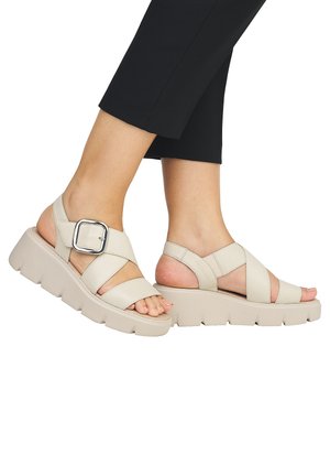 Beige platform sandals feature cross-strap design, large silver buckle accent, and thick, textured sole. Suitable for casual wear.