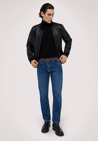 Chaqueta bomber de cuero negro sobre un suéter de cuello alto negro, combinada con jeans azules y botines negros. Modelo de pie mirando hacia adelante.