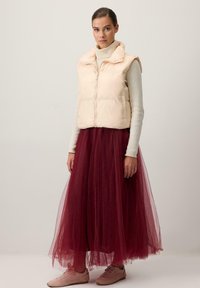 Világos rózsaszín, puffos mellény cipzárral az elején és magas gallérral, egy krémszínű pulóver és egy hosszú bordó tüllszoknya fölött viselve. Pink sportcipővel kombinálva.