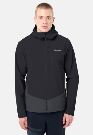 Giovane uomo che indossa una giacca con cappuccio nera e grigio scuro con cerniera frontale e tasca sul petto, in piedi contro uno sfondo chiaro e uniforme.