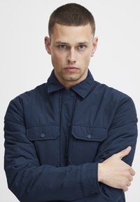 Tamsiai mėlynas paklotas švarkas su knopkių uždarymu, smailiu apykakle ir dviem krūtinės kišenėmis su anga uždarymo. Lygus paviršius ir struktūruota forma.