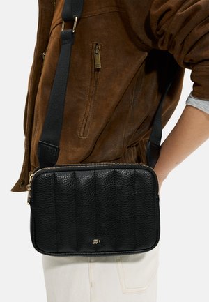 Czarna torebka typu crossbody o teksturowanej powierzchni z złotym zamkiem i małym logo, noszona na brązowej zamszowej kurtce oraz białych spodniach.