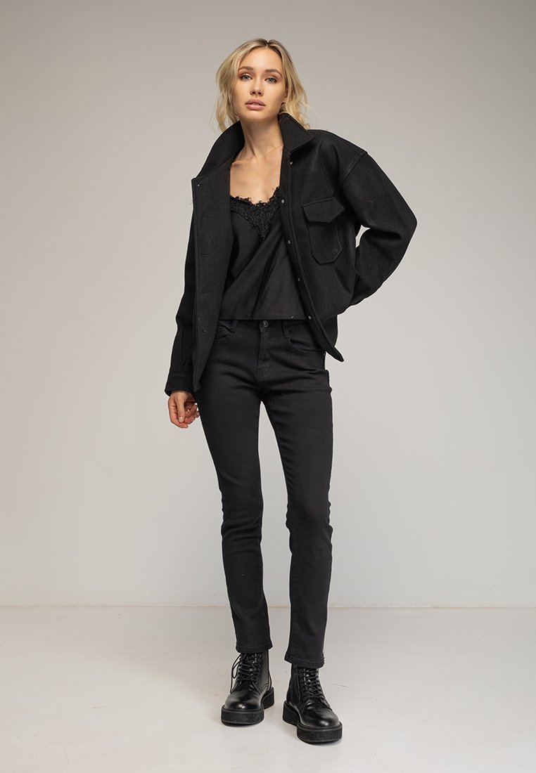 Chaqueta negra de gran tamaño con botones y grandes bolsillos, llevada sobre un top de encaje negro, combinada con jeans negros ajustados y botas negras con cordones.