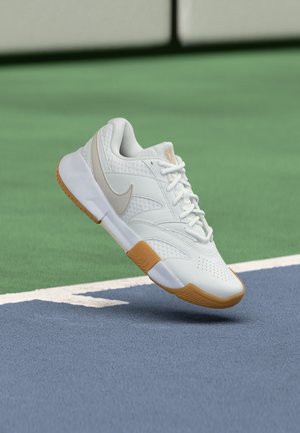 Weißer Sportschuh mit Gummisohle und beigem Nike-Swoosh, schwebt über einer blau-grünen Tennisplatzoberfläche.