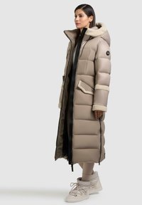 Cappotto imbottito lungo beige con colletto in ecopelliccia, chiusura a zip frontale, due tasche laterali e superficie testurizzata, mostrato insieme a calzature chiare.