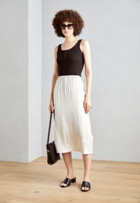 Top negro sin mangas con botones, falda midi blanca ligera, sandalias negras tipo slide y un bolso negro de hombro. Suelo de madera, fondo gris.