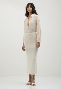 Chemise à rayures crème avec des manches bouffantes, associée à une jupe longue crème texturée et moulante, ainsi qu'à des mules ouvertes beige clair.