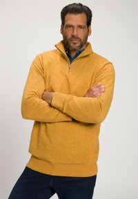 Gelber gestrickter Pullover mit einem Halbreißverschluss-Kragen, gerippten Bündchen und Saum. Der Pullover hat ein strukturiertes Muster und wird mit dunkelblauen Jeans kombiniert.